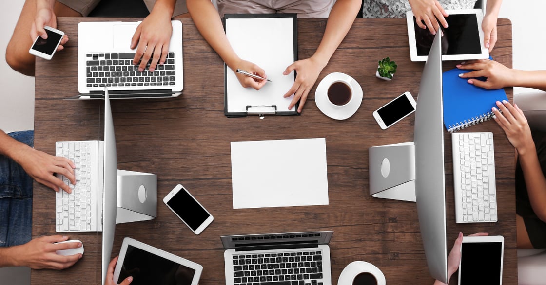 An overhead view of a table full of laptops, phones, tablets, and notepads. Several people's hands are visible as they work on various devices.